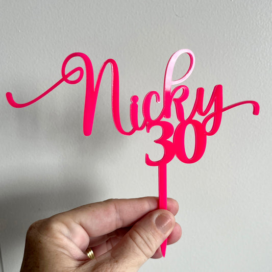 A bright pink acrylic cake topper with the text 'Nicky 30' in front of white background.