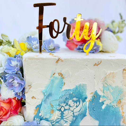 A cake topper with the word 'Forty' displayed in stylised lettering on a cake decorated with blue and gold icing and surrounded by colorful flowers.
