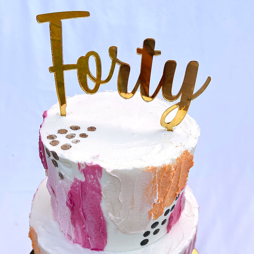 Close up of gold acrylic cake topper on a pink, orange and white cake.