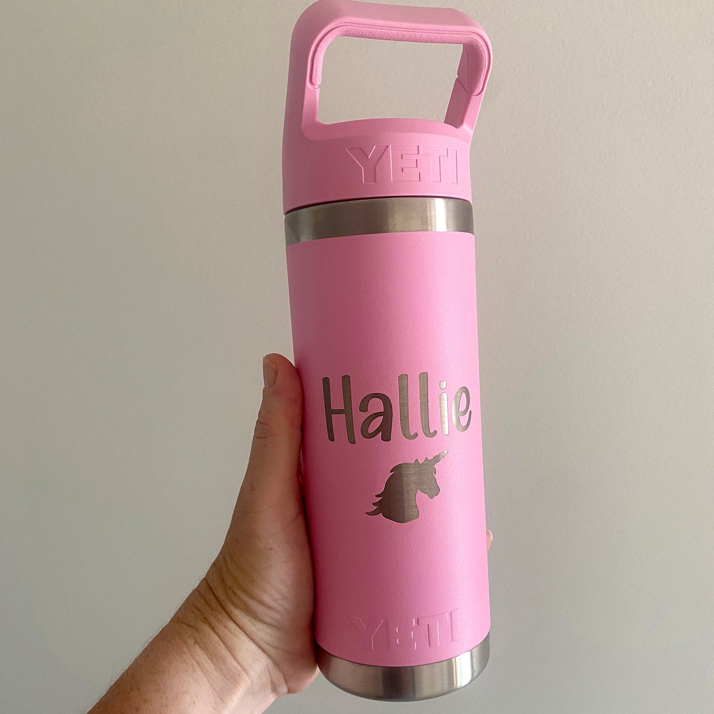 Pink YETI bottle with 'Hallie' and a unicorn design, held by a hand against a plain background.