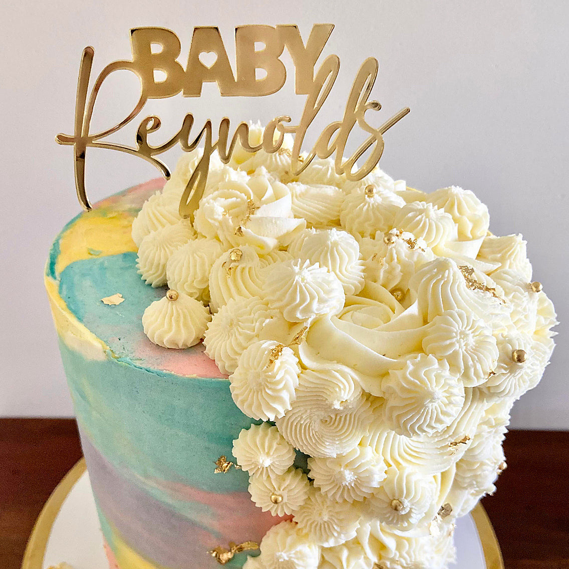 A gold-colored acrylic cake topper with the text 'BABY Reynolds' placed on a cake with white frosting.