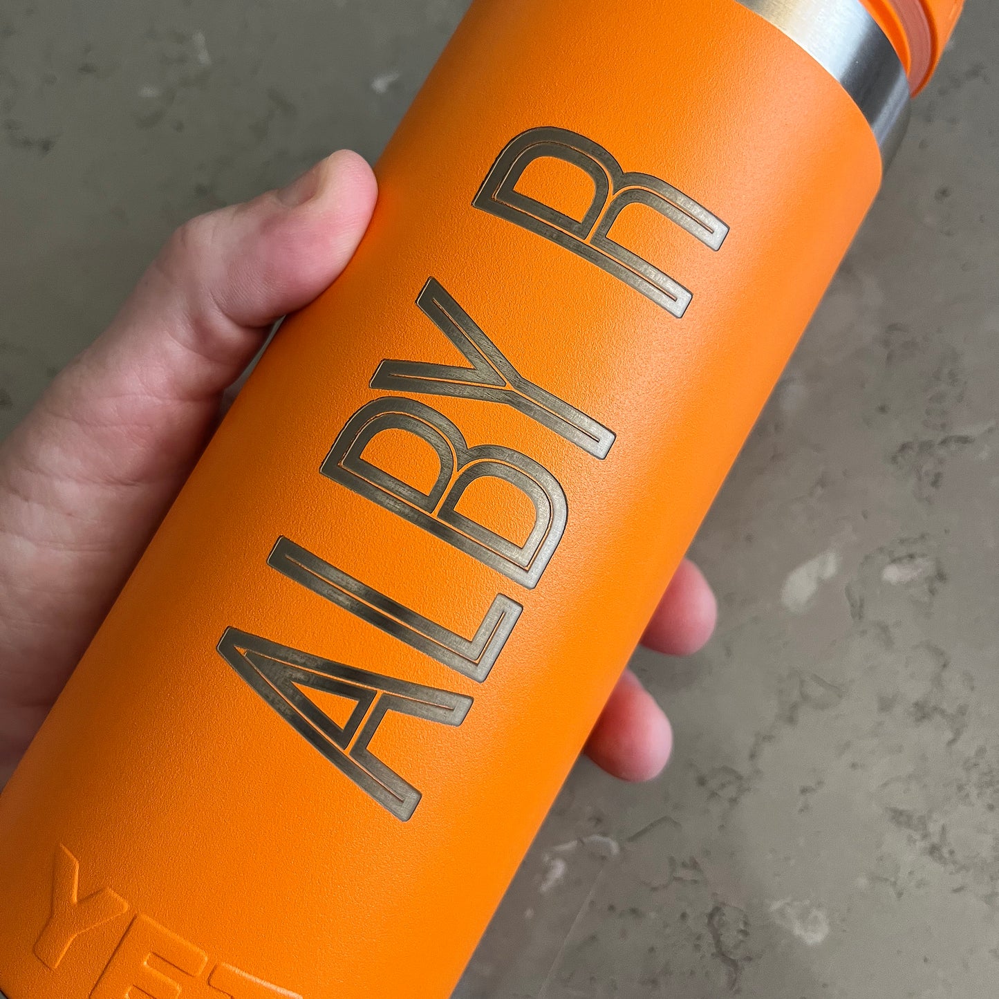 Orange water bottle with 'ALBY R' engraving held by a hand on a concrete surface.