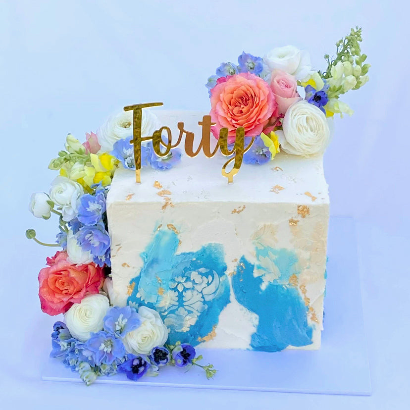 Simple script gold mirror cake topper on a white and blue cake, topper reads "Forty."