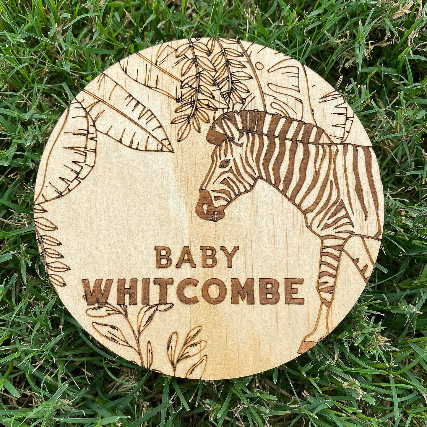 Wooden disc with zebra engraving and 'Baby Whitcombe' text on grass