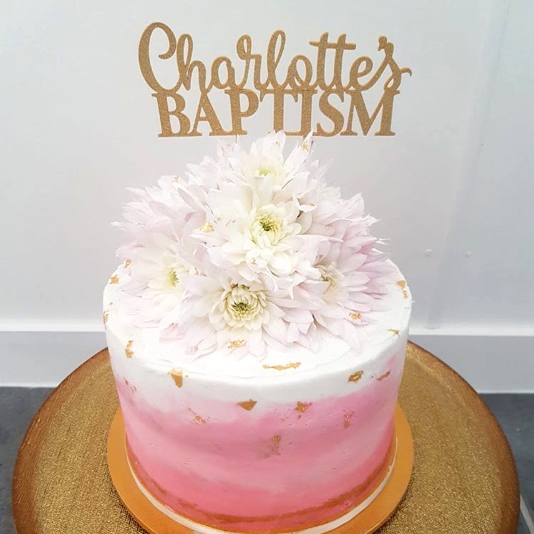 Gold glitter cake topper on a pink and white cake, reads "Charlotte's Baptism."